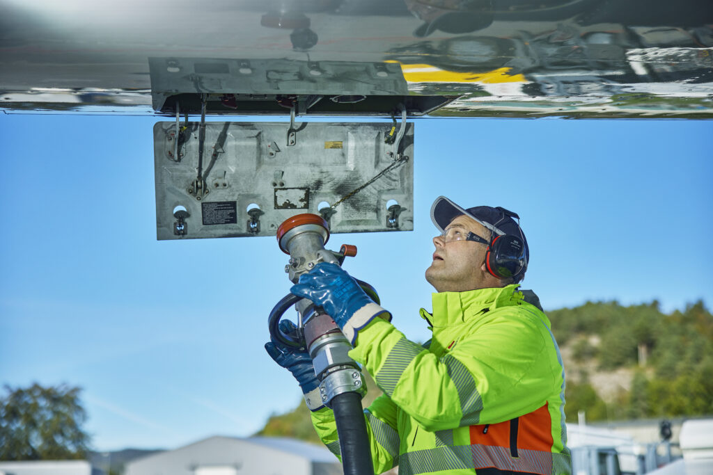 Mann i refleksvest, caps, vernebriller og hørselvern som løfter opp en slange med flydrivstoff mot flyet han skal fylle drivstoff på.