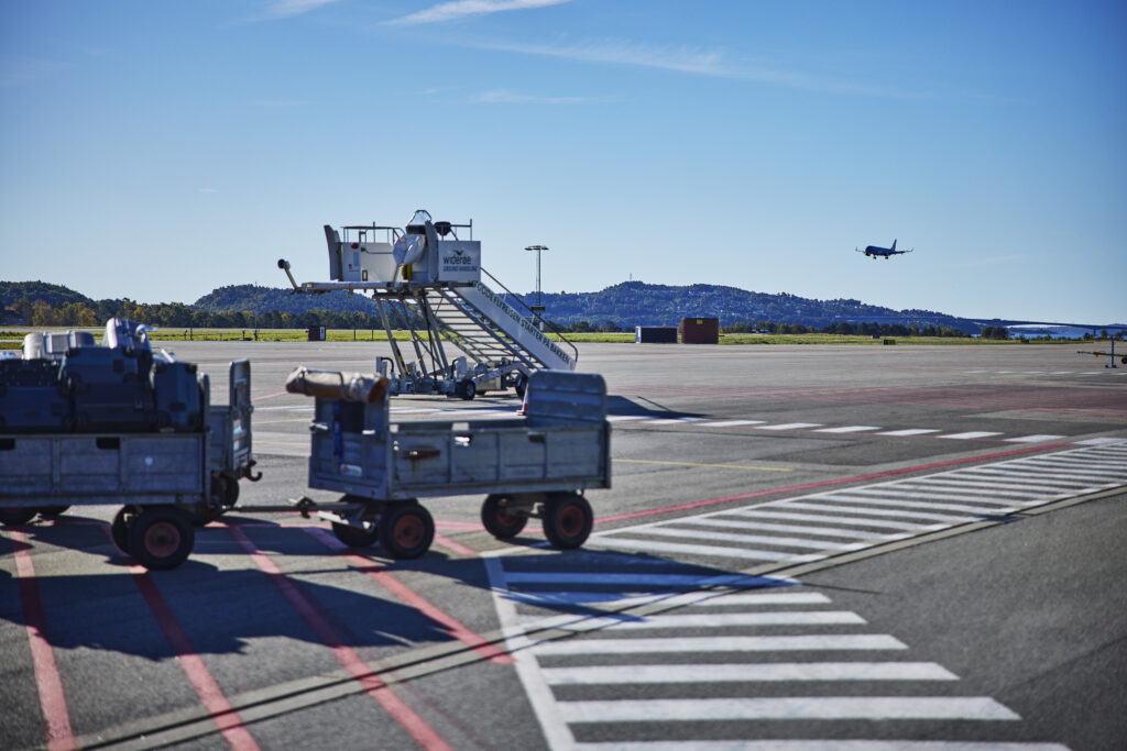 En flyplass med to hengere fylt av kofferter og annen bagasje. Ved horisonten ser vi et fly som har lettet og som reiser bort fra oss.