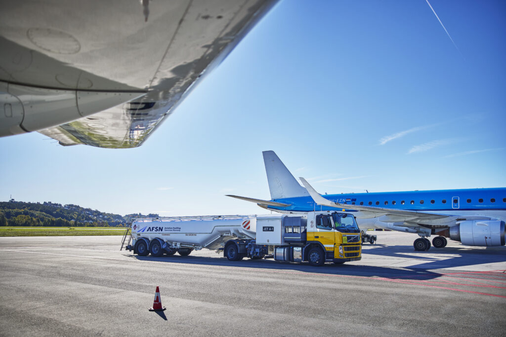 En flybane hvor man ser undersiden av en flyvinge og i midten av bildet ser man siden på en hvit og gul tankbil med AFSN logo på. Et hvitt og blått fly står bak tankbilen.