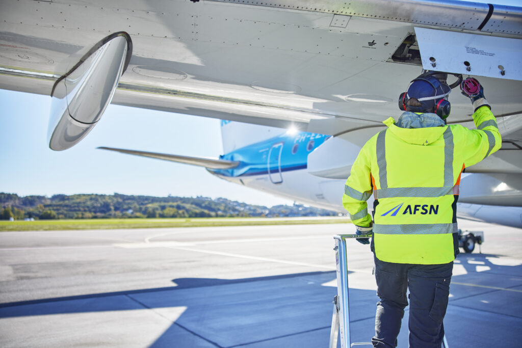 Mann med refleksvest og hørselvern som holder på med tanken på et fly. Han har ryggen til kamera hvor man ser en AFSN logo bakpå refleksvesten hans.