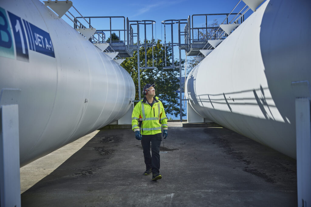 Mann i refleksvest og verneutstyr som står mellom to store tanker med drivstoff.